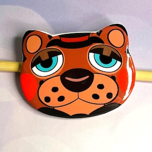 May include: A cartoon-style bear face with a brown body, black outlines, and a red blush on the cheeks. The bear has large, light blue eyes, a black nose, and a red stripe on its head. The design is glossy and appears to be a pin or badge.