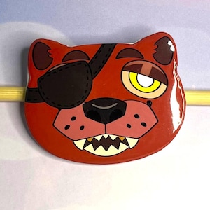 May include: A red, cartoon-style pin featuring a fox face with an eye patch, a yellow eye, and sharp teeth. The pin has a glossy finish and is likely a collectible or accessory.