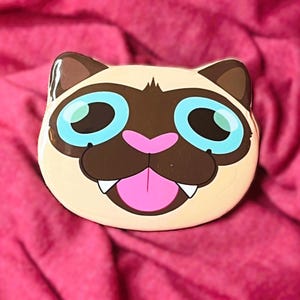 May include: A cat-shaped decorative item with a tan face, brown ears and muzzle, and large blue eyes. The cat has a pink nose and tongue, and white teeth. The item is set against a pink background.