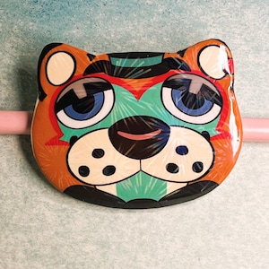 May include: A colorful, cartoon-style tiger face with large blue eyes, orange fur, and a black nose. The design includes green, red, and white accents. The tiger face is on a light pink stick, possibly a decorative item or a toy.