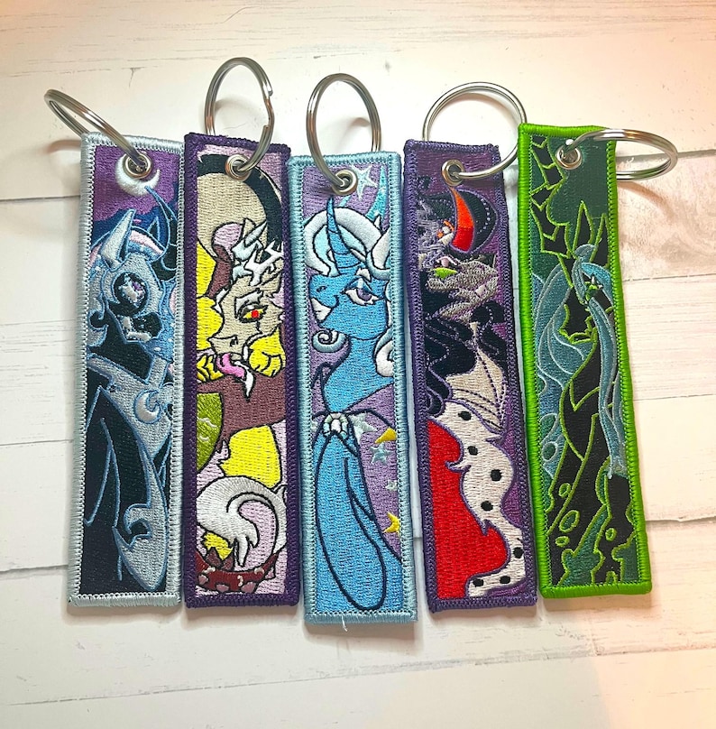 May include: Five embroidered keychains with silver key rings. Each keychain features a different cartoon character design in various colors, including blue, purple, green, yellow, and red. The keychains have a rectangular shape with a purple border.
