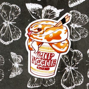 Könnte beinhalten: Ein Aufkleber mit einer Cartoon-Schlange in einer Tasse Nudeln. Die Schlange ist weiß und orange mit roten Augen. Die Nudeln befinden sich in einer gelben Tasse mit dem Text "CUP NOODLE" in Rot und "カッ プ ヌードル" in Schwarz.