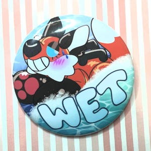 May include: A circular pin-back button featuring a stylized illustration of a black, orange, and white creature with blue eyes and pink cheeks. The creature is partially submerged in blue water with white foam, and the word WET is printed in large blue letters.