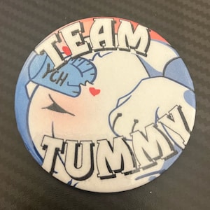 Puede incluir: Un botón blanco con un personaje de dibujos animados. El personaje es un animal blanco con ojos azules y un sombrero azul. El texto "TEAM TUMMY" está escrito en negro alrededor del personaje.