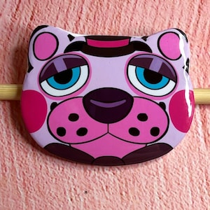 May include: A cartoon-style cat face pin with a glossy finish. The pin features a light purple base color with pink accents on the cheeks, ears, and nose. The eyes are blue with white highlights, and the nose is dark purple.