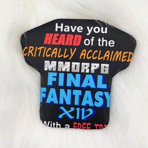 May include: A black t-shirt shaped magnet featuring white, red, orange, and blue text that reads: Have you HEARD of the CRITICALLY ACCLAIMED MMORPG FINAL FANTASY XIV With a FREE TRIAL.