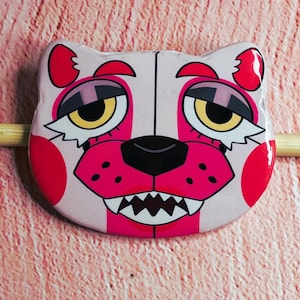 May include: A pink and white cartoon bear face button with red accents. The bear has yellow eyes, a black nose, and a wide, toothy grin. The button is on a light pink surface.