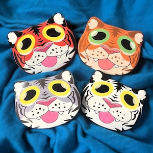 May include: Four decorative dishes in the shape of cat faces, each with a tiger-like design. The dishes feature yellow eyes, pink tongues, and are colored in red, orange, grey, and white. The background is a blue fabric.
