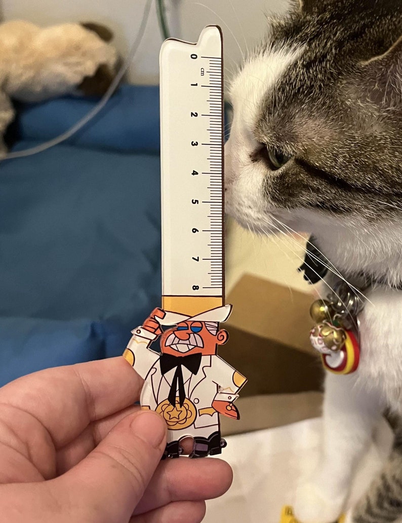 May include: A white and gray cat with a bell collar looks at a ruler with a cartoon character on it. The ruler is 8 cm long and has black markings. The cartoon character is wearing a white suit, a black bow tie, and a yellow hat. The character is holding a yellow star.