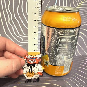 May include: A clear acrylic pin of a cartoon character wearing a white suit and a top hat. The character has a red nose, a mustache, and a pumpkin in his hand. The pin is next to a can of soda with a nutrition facts label. The label states that the soda contains 0 calories.