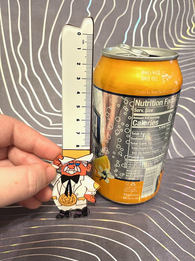 May include: A clear acrylic pin of a cartoon character wearing a white suit and a top hat. The character has a red nose, a mustache, and a pumpkin in his hand. The pin is next to a can of soda with a nutrition facts label. The label states that the soda contains 0 calories.