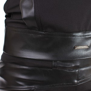 K&ouml;nnte beinhalten: Nahaufnahme eines schwarzen Outfits mit kontrastierenden Texturen. Das Design umfasst einen breiten Kunstleder-G&uuml;rtel mit einem kleinen Metallakzent. Das Outfit scheint aus einem Oberteil und einer Hose zu bestehen. Der G&uuml;rtel ist etwa 10 cm breit.
