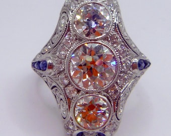May include: An antique silver ring with three large, round diamonds and smaller accent diamonds. The ring has a detailed filigree design and small blue gemstones. A classic piece of jewellery.