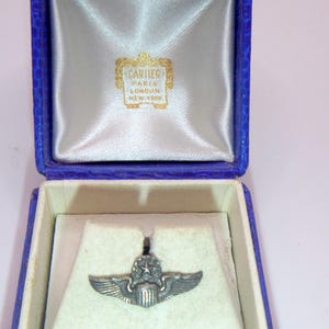 Puede incluir: Un broche plateado vintage con un diseño alado, presentado en una caja azul abierta con interior blanco. La caja tiene un exterior azul texturizado y una etiqueta que dice "CARTIER PARIS LONDON NEW YORK".