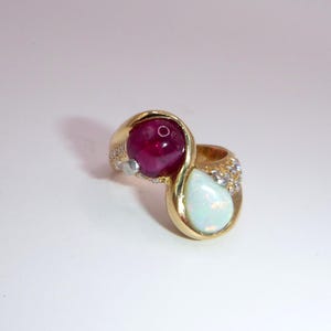 Può includere: Un anello d'oro con un grande rubino rosso scuro rotondo e una pietra bianca opalescente a forma di pera. L'anello ha un design a gambo diviso con piccole pietre di accento trasparenti. L'anello è su uno sfondo bianco.