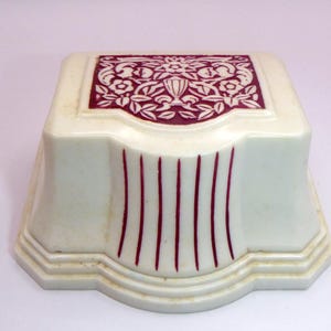 Op de afbeelding: Crèmekleurige doos in vintage-stijl met een decoratief bordeauxrood bloemmotief op de bovenkant. De doos heeft verticale bordeauxrode strepen op de voorkant en een getrapte basis. Het ontwerp doet denken aan de Art Deco-stijl.