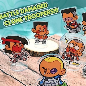 May include: Five cartoon character figurines of Star Wars Clone Troopers with battle damage. The figurines are made of clear acrylic and are displayed on a wooden surface. The text "BATTLE DAMAGED CLONE TROOPERS!!!" is visible in the image.