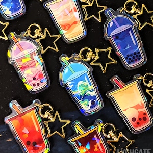 May include: A set of eight iridescent holographic charm keychains featuring different bubble tea flavors. Each keychain has a gold star charm attached to a gold chain. The keychains are arranged on a black surface.