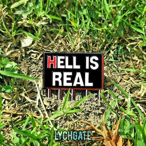May include: A black enamel pin with the words "HELL IS REAL" in white block letters, with the word "HELL" in red. The pin is rectangular with a red border and silver pin backs. The pin is set against a background of green grass and brown earth.
