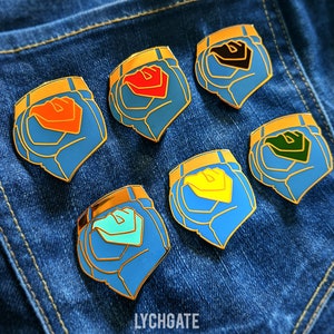 May include: Six enamel pins featuring a blue, gold and white design with different coloured accents. The pins are arranged in a row on a blue denim background. The pins feature a stylised design of a pair of jeans.