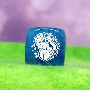 May include: A blue dice with a white illustration of a cartoon character with a star-shaped halo. The dice is on a green surface.