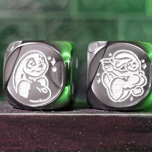 May include: Two square, metallic dice with engraved cartoon characters. One die features a bunny, the other an angel. The dice are displayed on a dark, rectangular base, with a blurred background of decorative figurines.