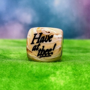 Może przedstawiać: Biała i brązowa kostka do gry z napisem "Have at thee!" napisanym czarnym kolorem.