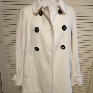 May include: Cream-colored double-breasted pea coat with black buttons. The coat has a classic design with a notched collar, front pockets, and folded cuffs. The coat is displayed on a mannequin.