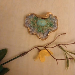 May include: A gold-toned brooch featuring a turquoise cabochon stone surrounded by clear and green glass beads.