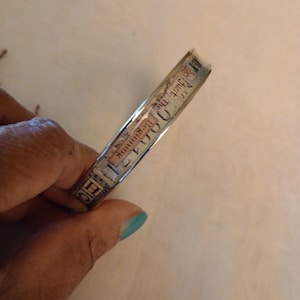 Puede incluir: Una pulsera de metal plateado con un diseño de papel que presenta las palabras "The Juice" y "Delicious" en una fuente de estilo vintage.