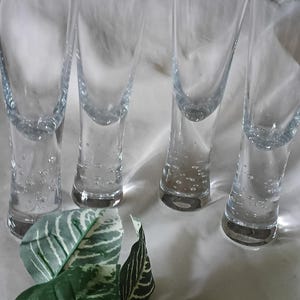 May include: Four clear glass shot glasses with rounded bottoms. The glasses are tall and slender, with a slight taper towards the top. The shot glasses are arranged in a row. Green and white leaves are in the foreground.