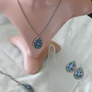 Puede incluir: Un conjunto de joyas plateadas que incluye un collar, una pulsera y pendientes. Cada pieza tiene un colgante o dije en forma de l&aacute;grima con un dise&ntilde;o de mosaico en tonos azules, rosas y verdes. El collar y la pulsera tienen eslabones de cadena.