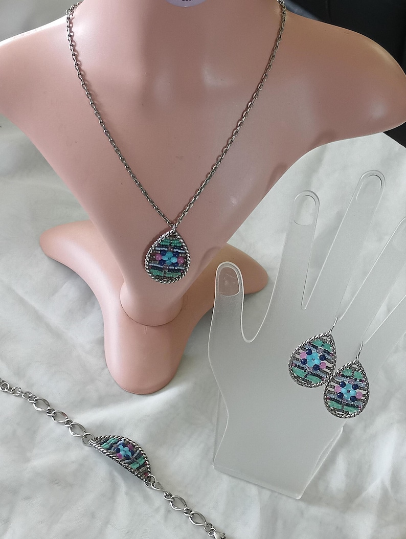 Puede incluir: Un conjunto de joyas plateadas que incluye un collar, una pulsera y pendientes. Cada pieza tiene un colgante o dije en forma de l&aacute;grima con un dise&ntilde;o de mosaico en tonos azules, rosas y verdes. El collar y la pulsera tienen eslabones de cadena.