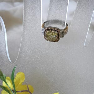 Puede incluir: Un anillo dorado y plateado con una piedra preciosa cuadrada de color amarillo claro. El anillo tiene una banda retorcida y un engaste decorativo con pequeñas piedras transparentes. El anillo se exhibe en un soporte transparente en forma de mano.