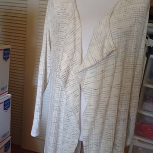 May include: A long white cardigan with a black and white striped pattern. The cardigan has a V-neck and long sleeves.
