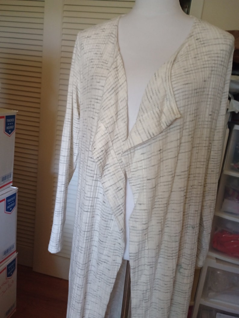 May include: A long white cardigan with a black and white striped pattern. The cardigan has a V-neck and long sleeves.