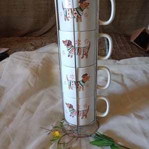 May include: Set of four white ceramic mugs with a dachshund wearing a Christmas sweater design. The mugs are stacked in a metal stand.