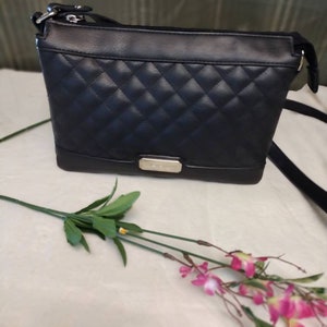 May include: A black quilted crossbody bag with a silver zipper and a silver nameplate. The bag is on a white surface with a pink flower.