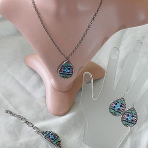 Puede incluir: Un conjunto de joyas plateadas que incluye un collar, una pulsera y pendientes. Cada pieza tiene un colgante o dije en forma de l&aacute;grima con un dise&ntilde;o de mosaico en tonos azules, rosas y verdes. El conjunto se exhibe en un maniqu&iacute; y un expositor en forma de mano.