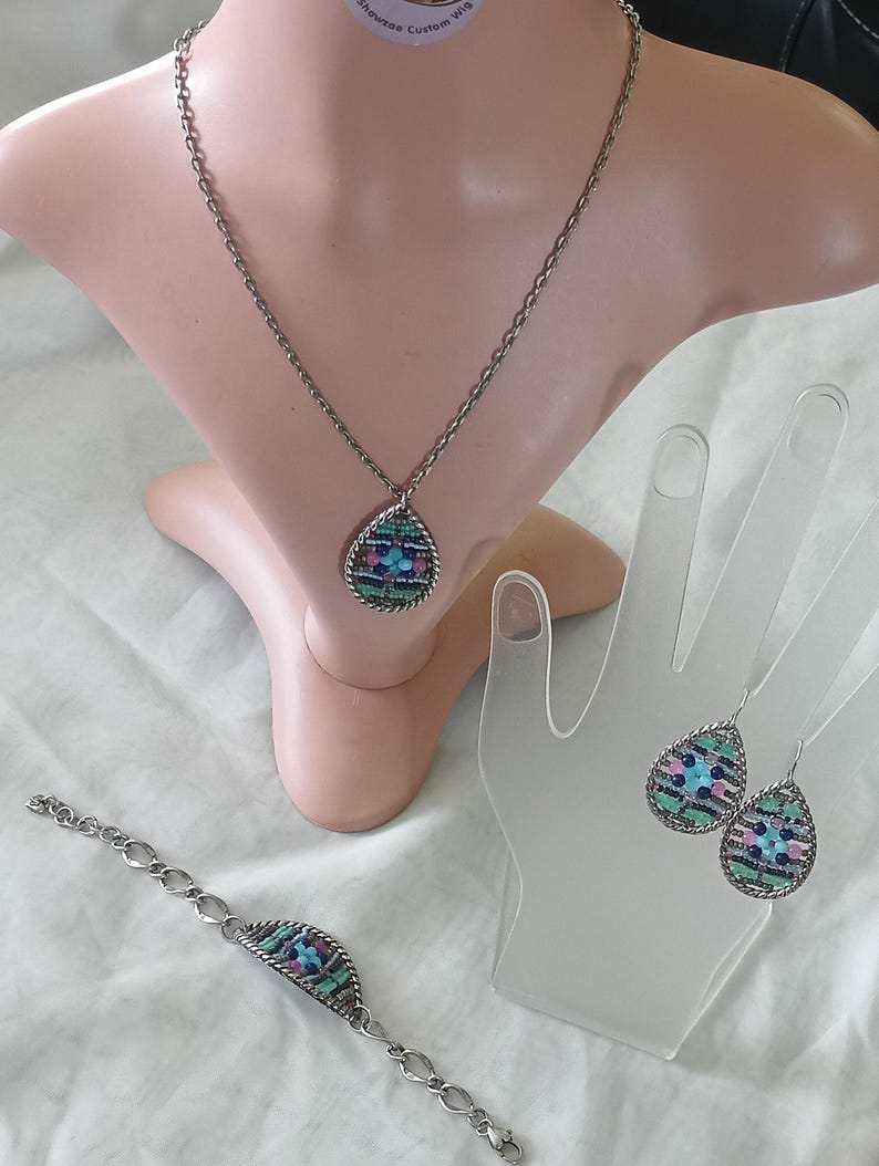 Puede incluir: Un conjunto de joyas plateadas que incluye un collar, una pulsera y pendientes. Cada pieza tiene un colgante o dije en forma de l&aacute;grima con un dise&ntilde;o de mosaico en tonos azules, rosas y verdes. El conjunto se exhibe en un maniqu&iacute; y un expositor en forma de mano.