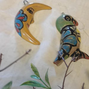 May include: Two decorative ornaments: a crescent moon and a bird. The moon is yellow with a painted face and blue and red floral accents. The bird is green, blue, and yellow with black details. Both have silver hanging loops.