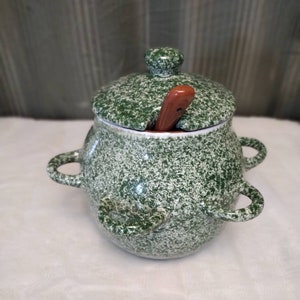 Könnte beinhalten: Grüne Keramik-Zuckerdose mit Deckel und Holzlöffel. Die Schüssel hat eine gesprenkelte grün-weiße Glasur.