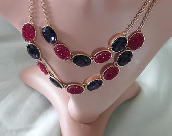 May include: A gold-toned necklace with two strands, each featuring oval-shaped gemstones. The gemstones alternate between a deep red and a dark purple hue. The necklace is displayed on a mannequin.