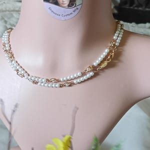 Puede incluir: Un collar de dos hilos con pequeñas perlas blancas y eslabones de cadena dorados. El collar se exhibe en un maniquí. Una pegatina con el texto "Showzae Custom Wig" es visible.