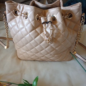 Può includere: Una borsa a secchiello trapuntata beige con una tracolla a catena dorata e una chiusura a cordoncino dorata.