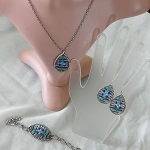 Puede incluir: Un conjunto de joyas plateadas que incluye un collar, una pulsera y pendientes. Cada pieza tiene un colgante o dije en forma de l&aacute;grima con un dise&ntilde;o de mosaico en tonos azules, rosas y verdes. Las joyas se exhiben en un maniqu&iacute; y una mano.