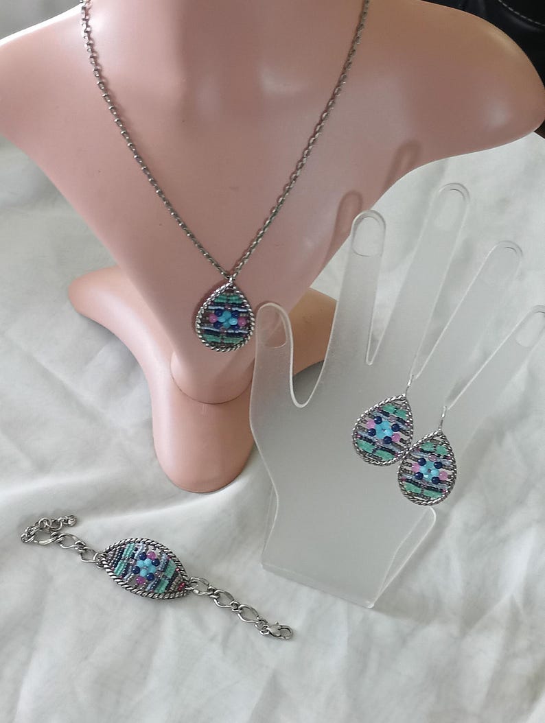 Puede incluir: Un conjunto de joyas plateadas que incluye un collar, una pulsera y pendientes. Cada pieza tiene un colgante o dije en forma de l&aacute;grima con un dise&ntilde;o de mosaico en tonos azules, rosas y verdes. Las joyas se exhiben en un maniqu&iacute; y una mano.