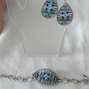 Puede incluir: Un conjunto de joyas con pendientes de l&aacute;grima y una pulsera. Los pendientes y la pulsera tienen un marco de metal plateado con un dise&ntilde;o tejido de cuentas azules, rosas y verdes. La pulsera tiene una cadena con eslabones ovalados.