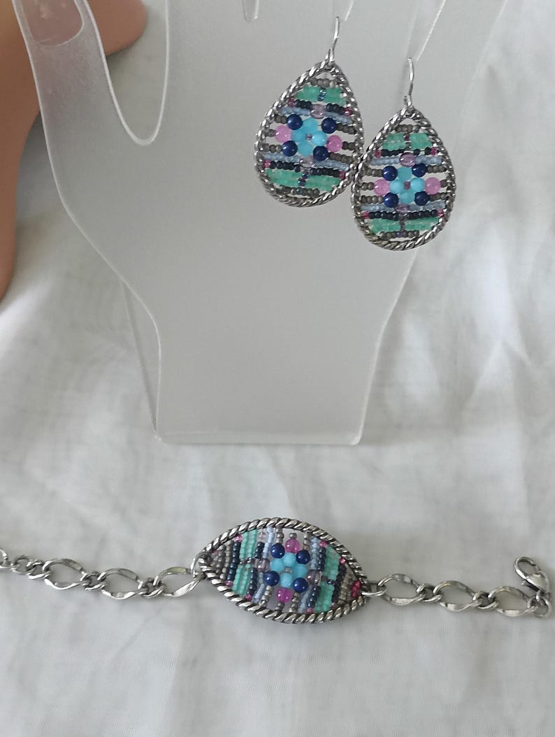 Puede incluir: Un conjunto de joyas con pendientes de l&aacute;grima y una pulsera. Los pendientes y la pulsera tienen un marco de metal plateado con un dise&ntilde;o tejido de cuentas azules, rosas y verdes. La pulsera tiene una cadena con eslabones ovalados.
