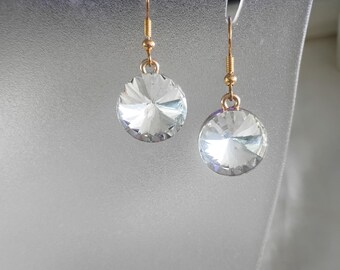 May include: A pair of clear, round crystal earrings with gold-colored hooks. The earrings are faceted, reflecting light. The jewelry is displayed on a clear, curved surface.
